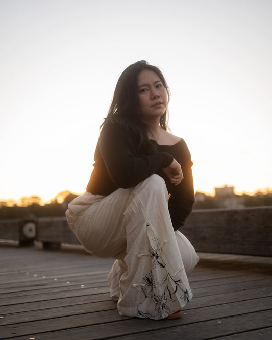 A woman crouching on a wooden boardwalk at sunset, wearing a black long-sleeve top and loose white pants with black floral embroidery.