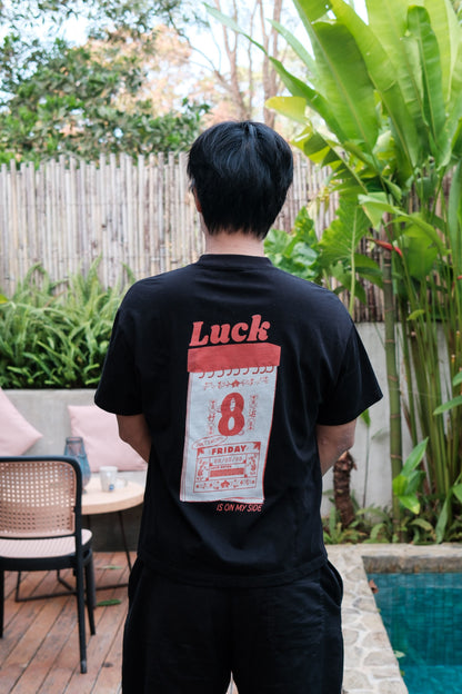 Man wearing a black t-shirt with Luck 8 graphics, standing outdoors near a pool and garden.