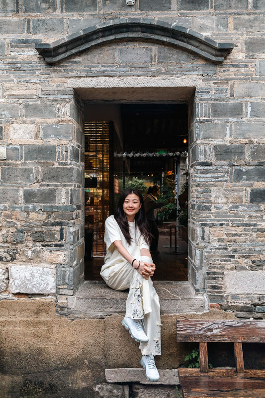 A woman sitting on the stone steps of a rustic gray brick building doorway, wearing a light beige short-sleeve top and matching pants with white sneakers.