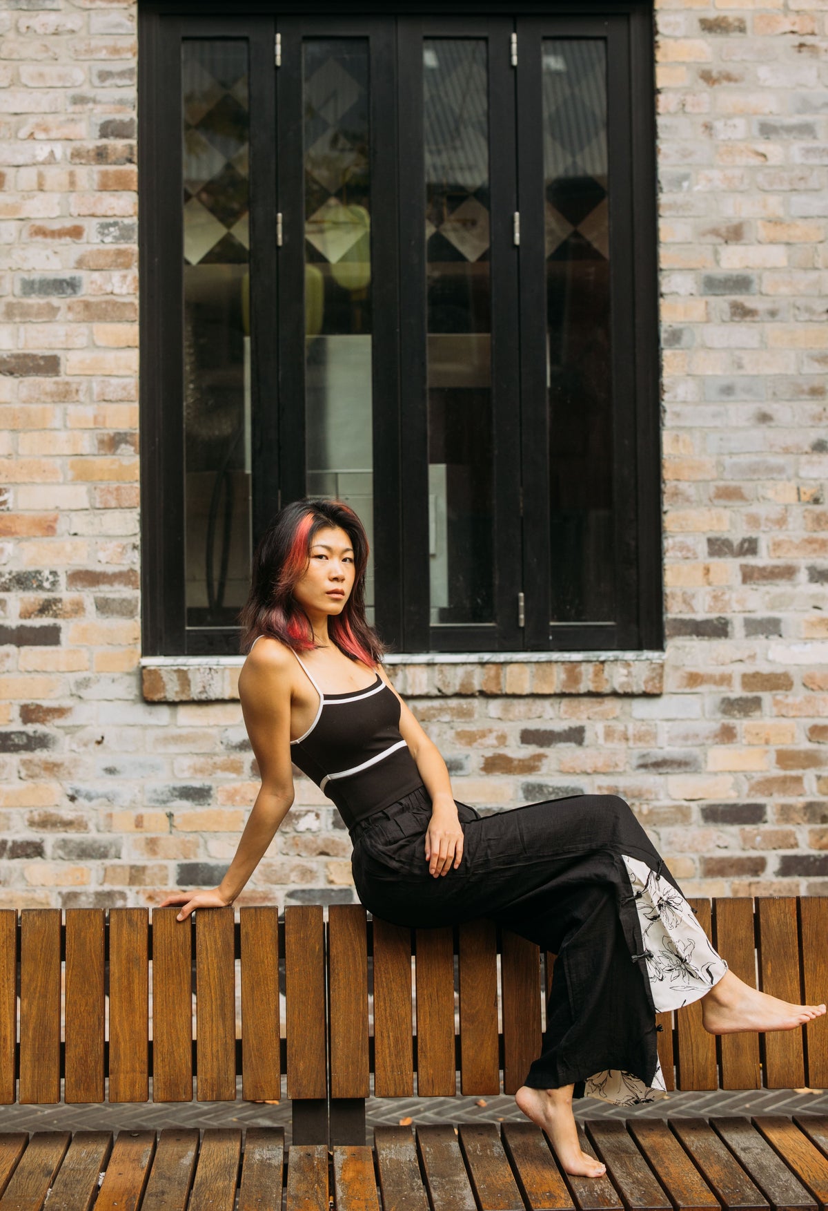 A woman with pink-tinted hair sits barefoot on a wooden bench against a brick wall, wearing a black pants with patterned print on the lower leg.