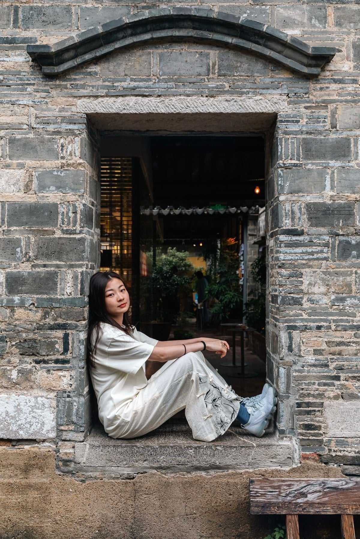 A woman sits on the stone ledge of an old brick building window, wearing a cream-colored short-sleeve top and matching pants with light blue sneakers.