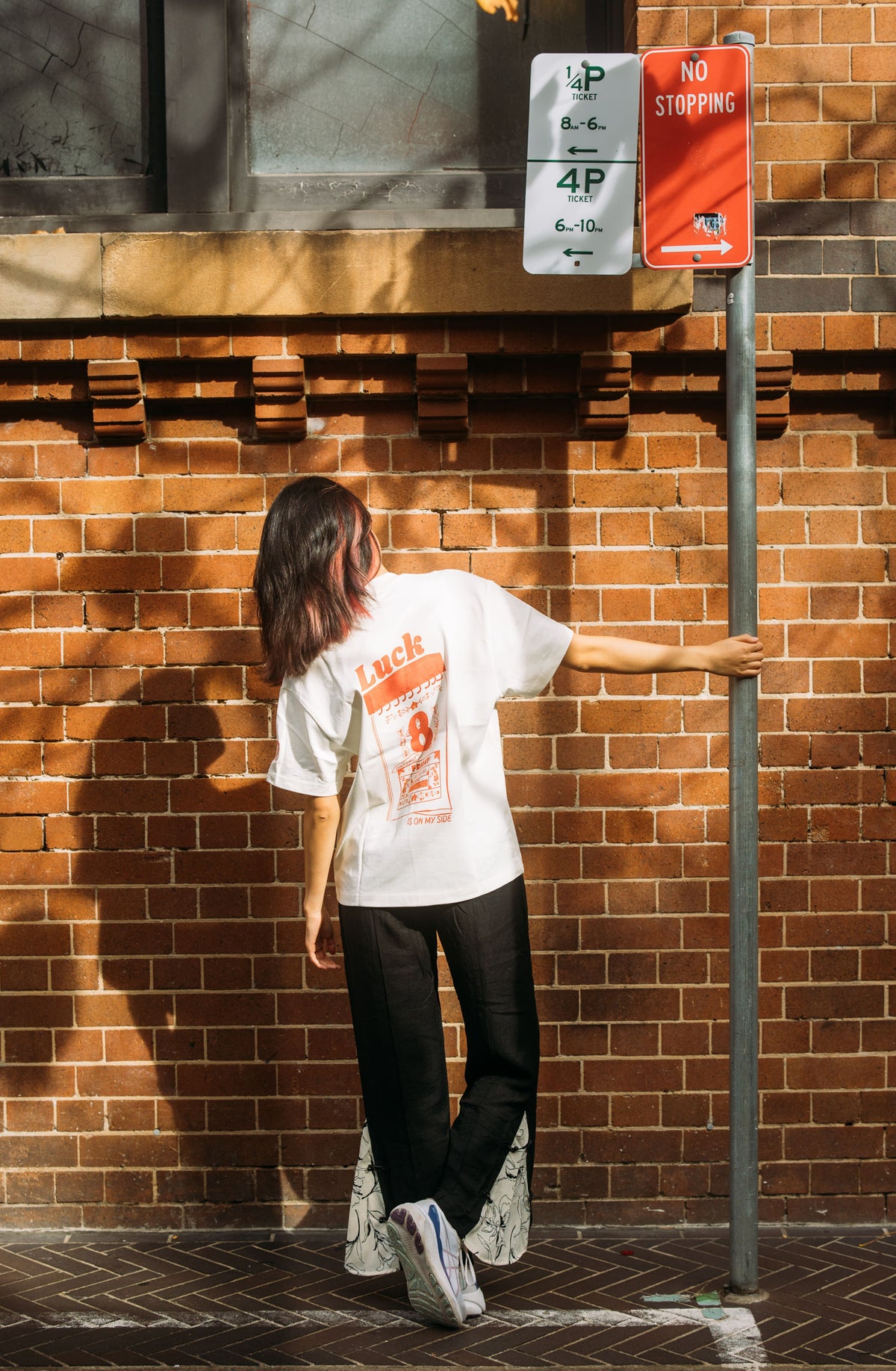 A person stands with their back to the camera against a red brick wall, wearing a white graphic t-shirt with Luck 8 design, black pants, and white sneakers, while holding onto a metal street sign pole.