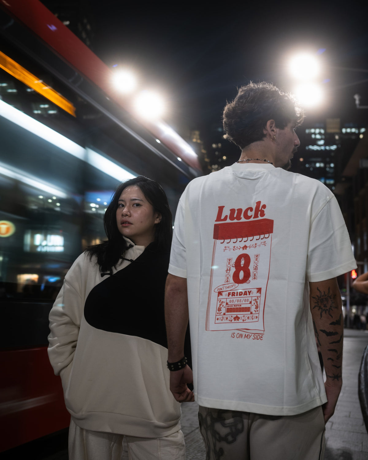 A man and a woman in an urban setting. The man is wearing a white t-shirt with a large red Luck 8 graphic print on the back and the woman wears a Yin Yang Sweater