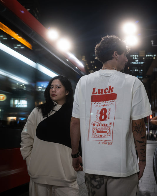 A man and a woman in an urban setting. The man is wearing a white t-shirt with a large red Luck 8 graphic print on the back and the woman wears a Yin Yang Sweater