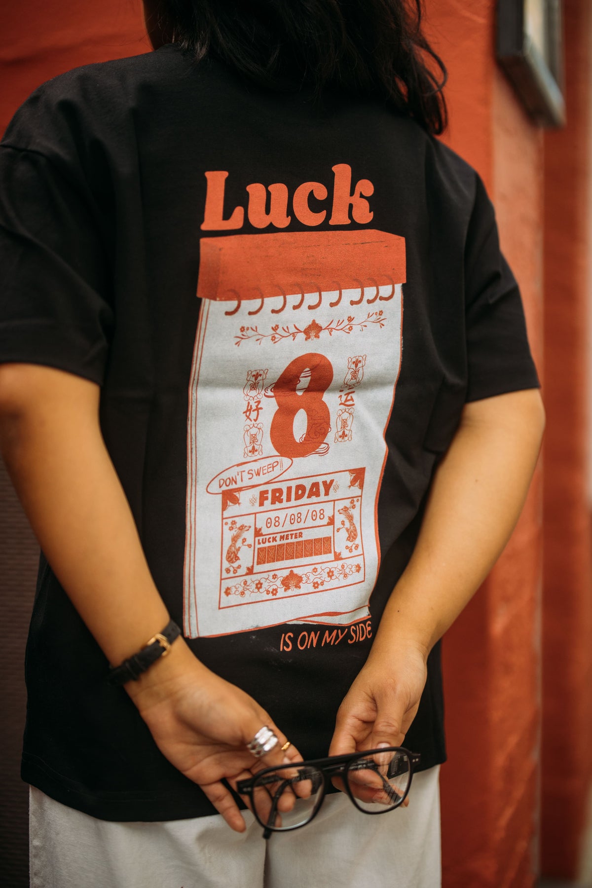 A person wearing a black t-shirt with a large graphic print on the back. The graphic features the word 'Luck' in red at the top, a white rectangular panel with a red '8' and vintage-style red calendar illustrations.