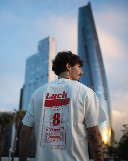 A man wearing a white t-shirt with a Luck 8 graphic print on the back, standing in front of modern skyscrapers at sunset.