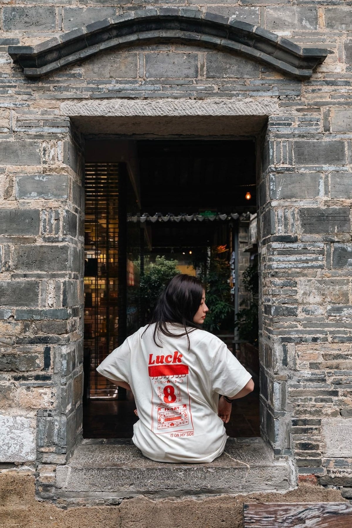A woman sitting in a stone doorway wearing a white t-shirt with a Luck 8 graphic print on the back, set against a rustic gray brick wall.