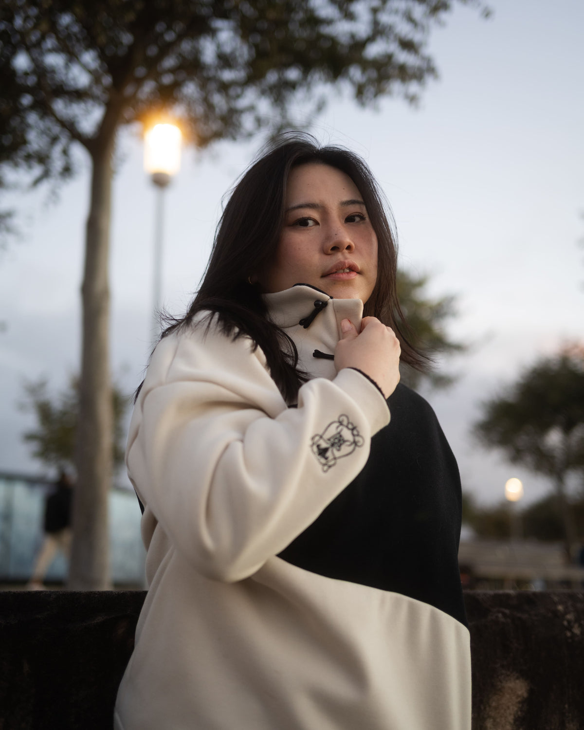 A woman wearing a color-blocked sweatshirt with a cream and black design, standing outdoors in a park-like setting with trees and streetlights in the background.